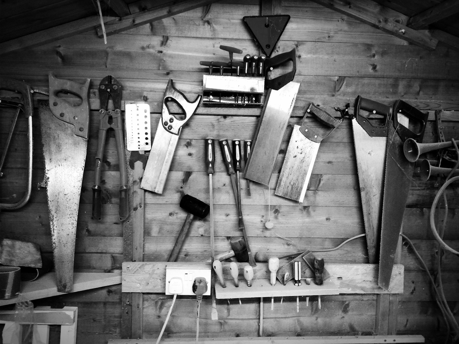 A black and white photograph of hand tools hung in rows on the plank wall of a workshop shed, saws, chisels and a mallet arranged above a bench.