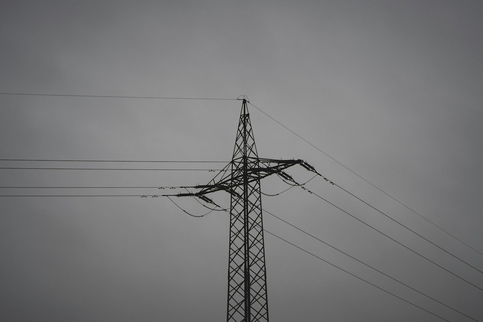 A black electric utility pole and power lines against a uniform gray sky, minimal and monochromatic, high negative space above the lines.