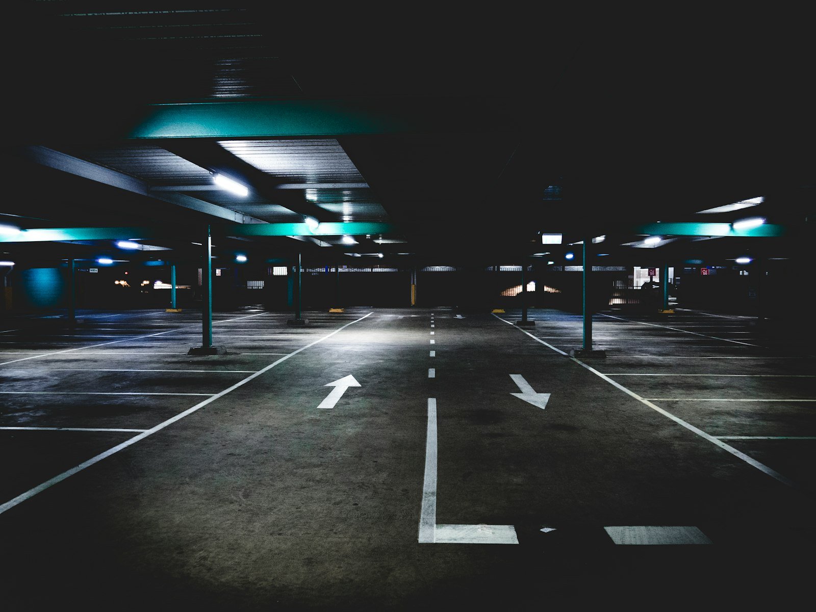 A dark, empty multi-storey car park with white directional arrows painted on the concrete floor and rows of vacant bays under strip lighting.