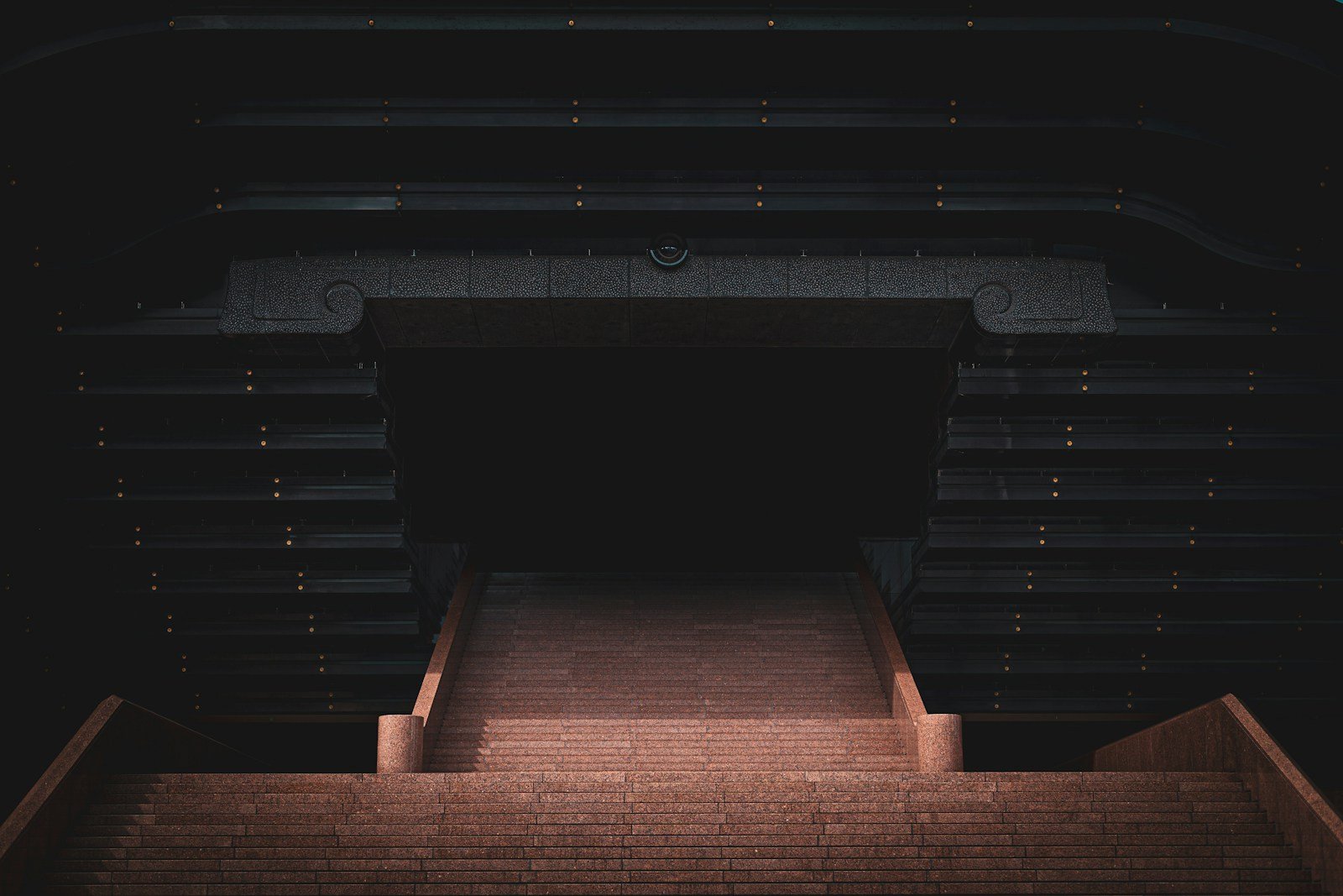Stone stairs illuminated from below leading upward to a pure black void, warm brick light fading at the threshold of darkness.