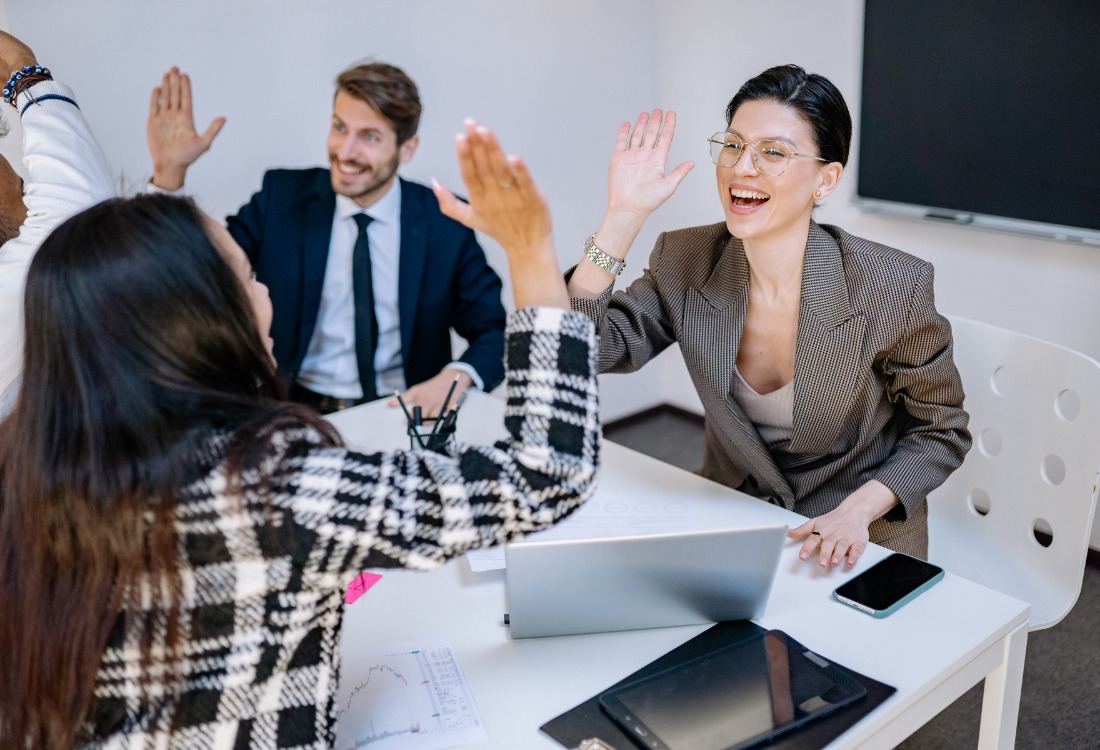Four colleagues in a modern office space, smiling and giving each other high fives across a table with a laptop, tablet, and smartphone on it, celebrating a successful outcome or team achievement.