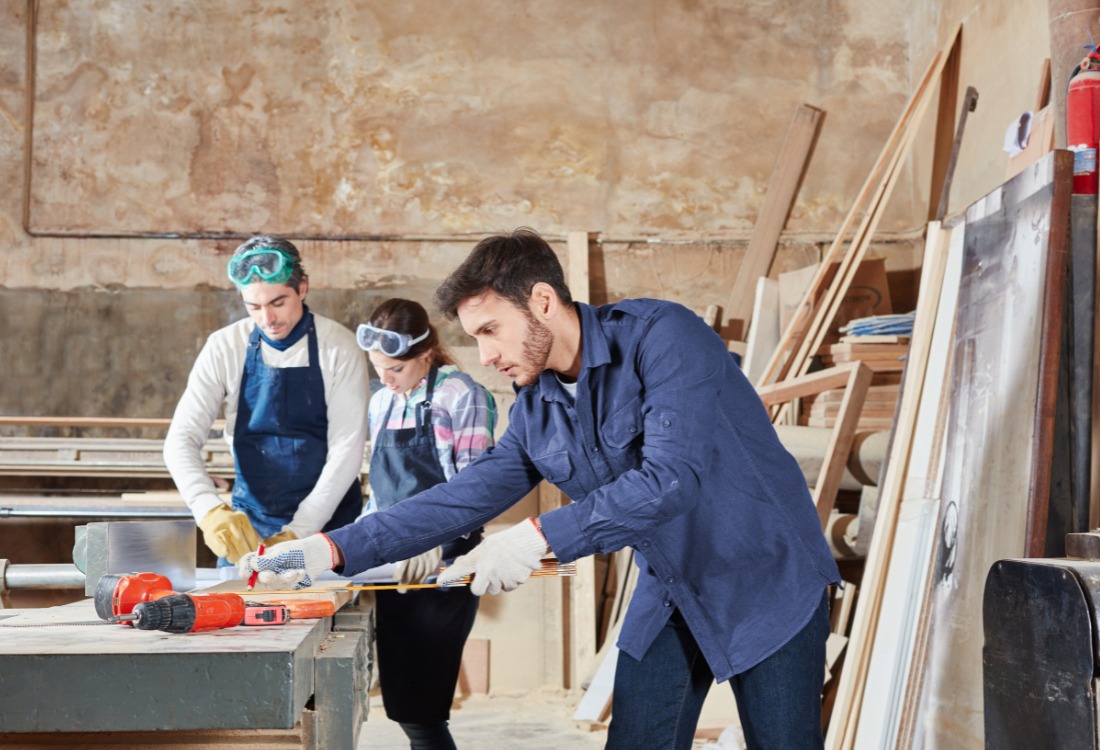 An apprentice measuring and marking a wooden board in a workshop, preparing for precise cuts, exemplifying foundational skills in carpentry and joinery.