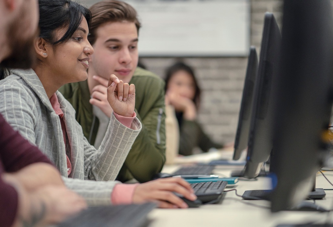 A focused group of apprentices engaged in a commercial training course, with one of the students animatedly explaining something on a computer screen while the other observes intently.