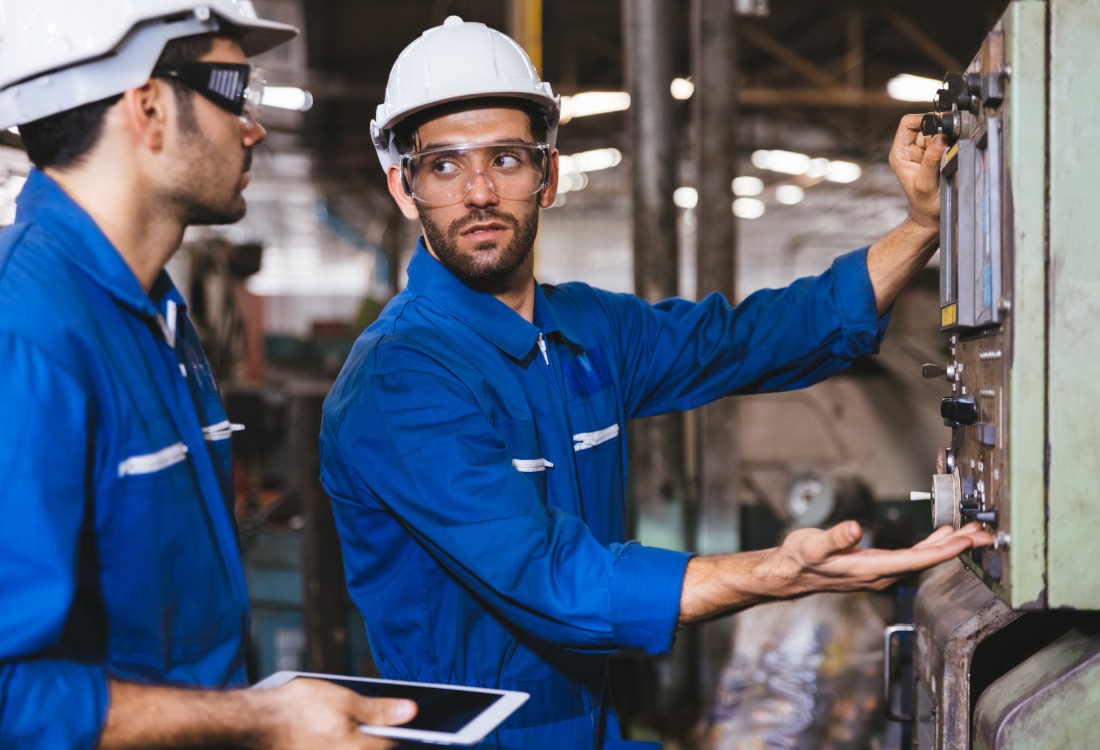 Two maintenance technician apprentices in blue overalls inspecting and troubleshooting industrial machinery, demonstrating hands-on training in a Maintenance Technician Apprenticeship.