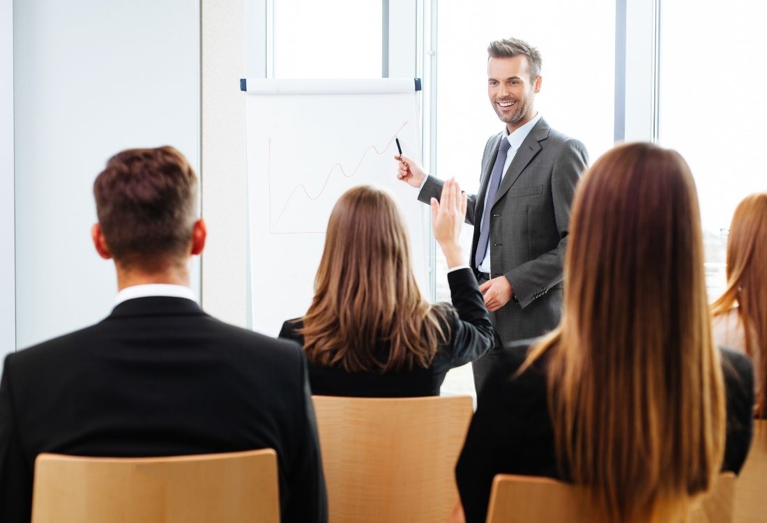 A male trainer presenting to a group during a leadership training session, with a female participant raising her hand to ask a question, illustrating interactive learning and engagement.