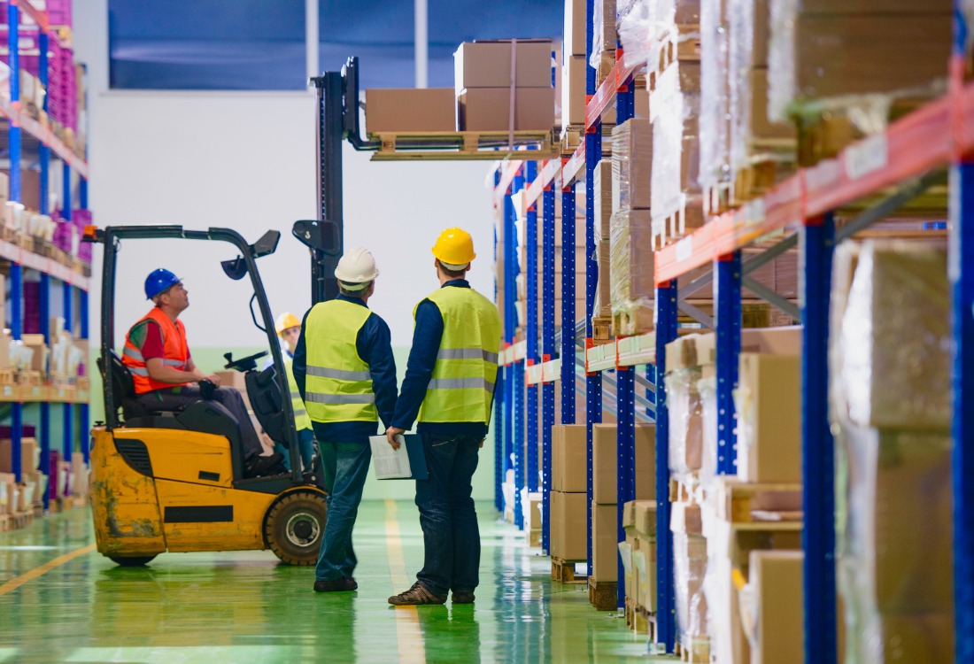 A forklift operator navigating a warehouse aisle, with three colleagues observing, illustrating hands-on training in a certified forklift operator course.