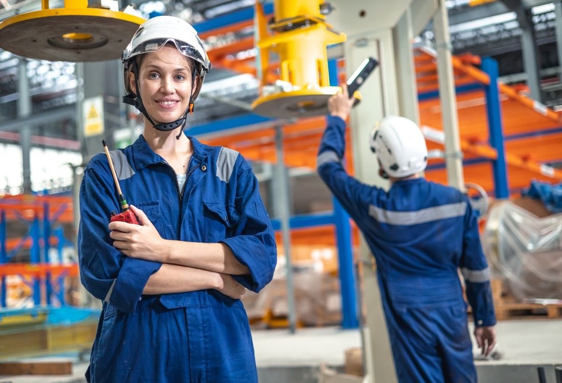 A female rail engineer wearing a white hard hat and blue overalls smiles at the camera, holding a two-way radio. In the background, another person in similar attire works with machinery.
