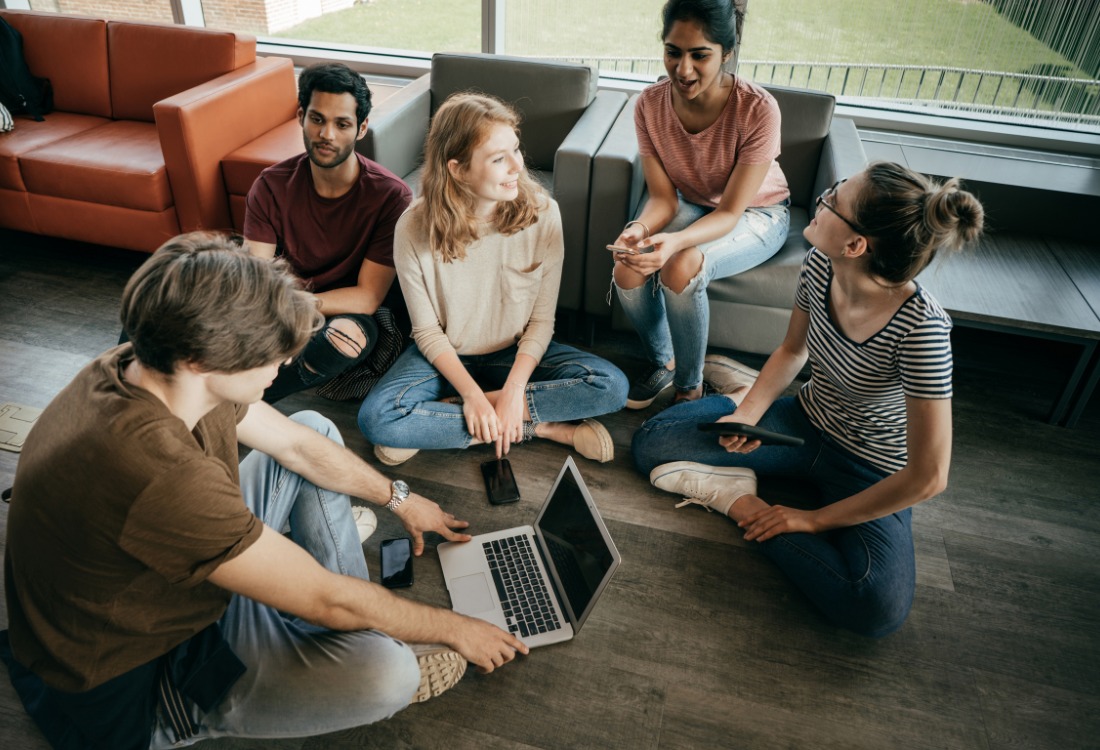 A group of international students seated on the floor discussing UK residency criteria, with one student using a laptop.