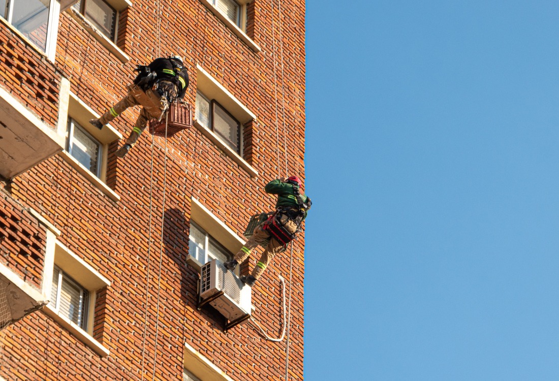 Workers using ropes and safety harnesses to perform maintenance at height on a tall brick building, demonstrating the need for safe bespoke access solutions.