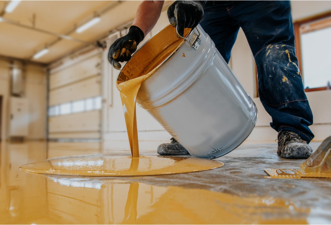 Worker pouring liquid resin onto an industrial floor during installation.