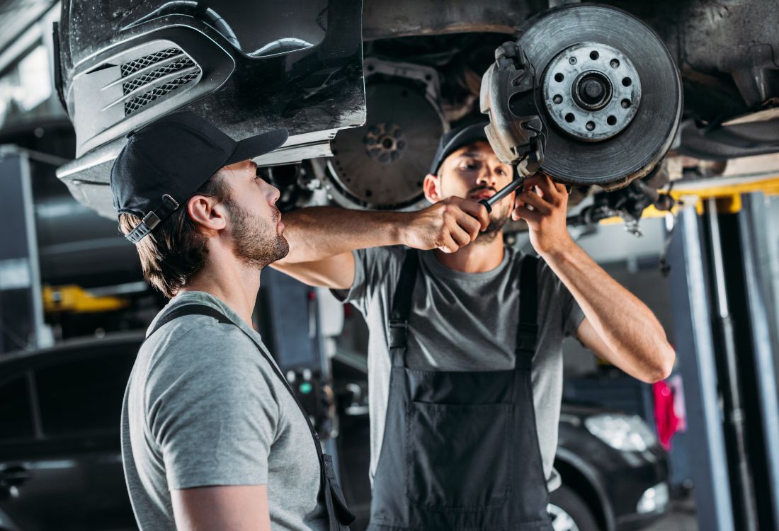 Two auto mechanics working under a raised vehicle in a garage, focusing on repairing the brake system while wearing caps and work overalls.