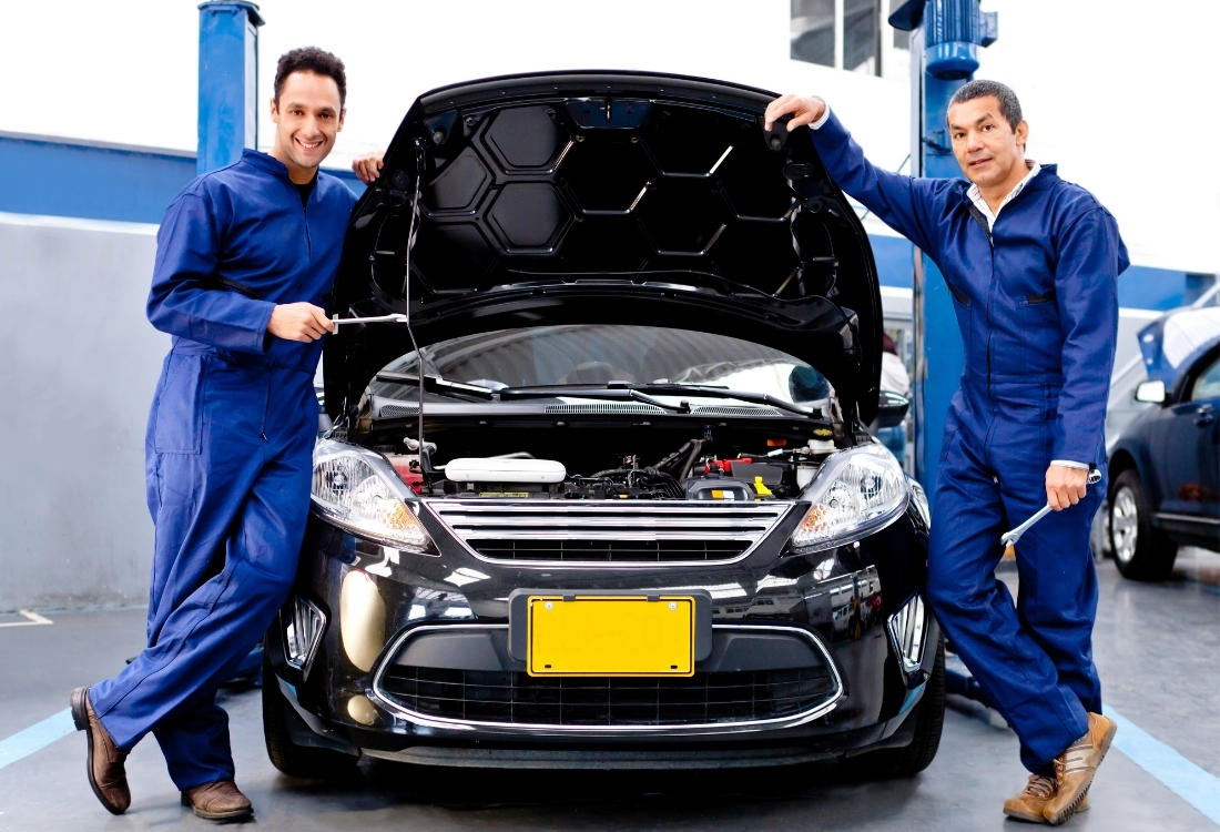 Two smiling auto mechanics in blue overalls standing beside a black car with its hood open inside a garage, holding tools and posing confidently.