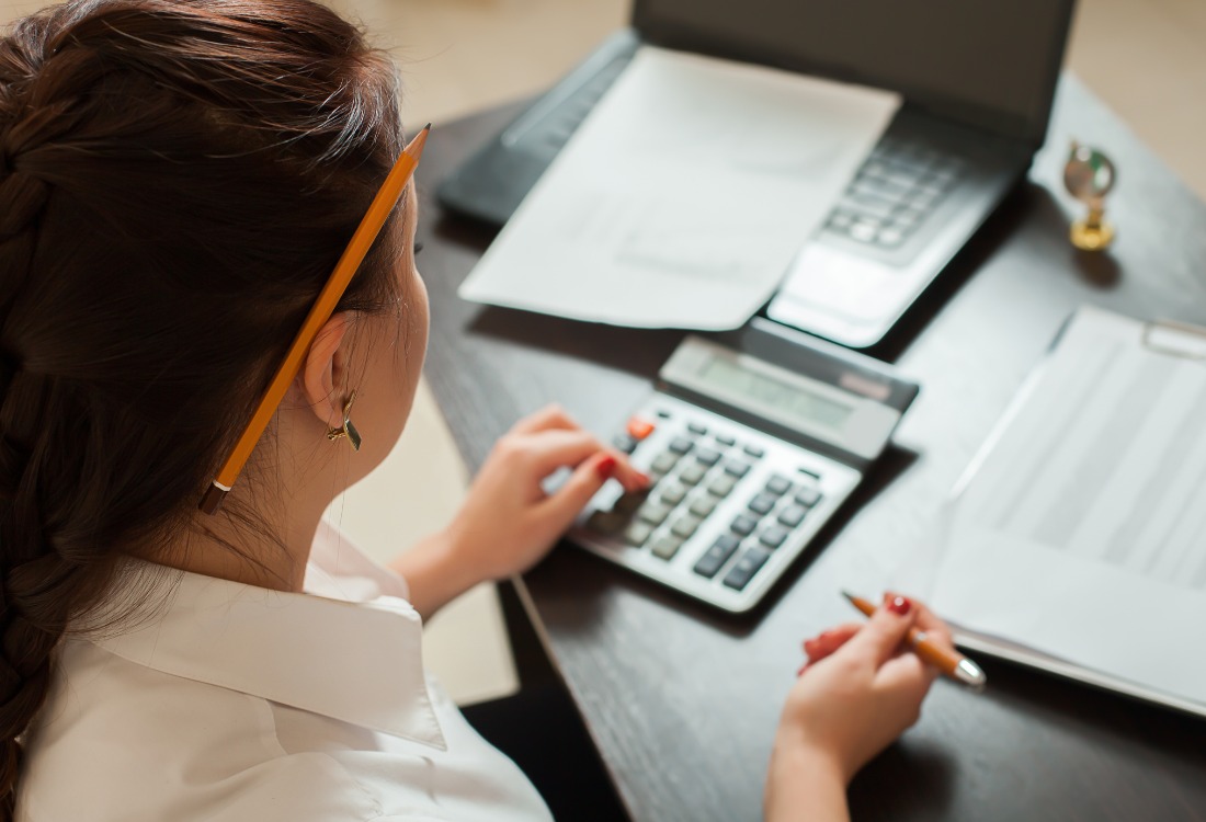 Bookkeeper reviewing financial records and using calculator at desk with documents. 