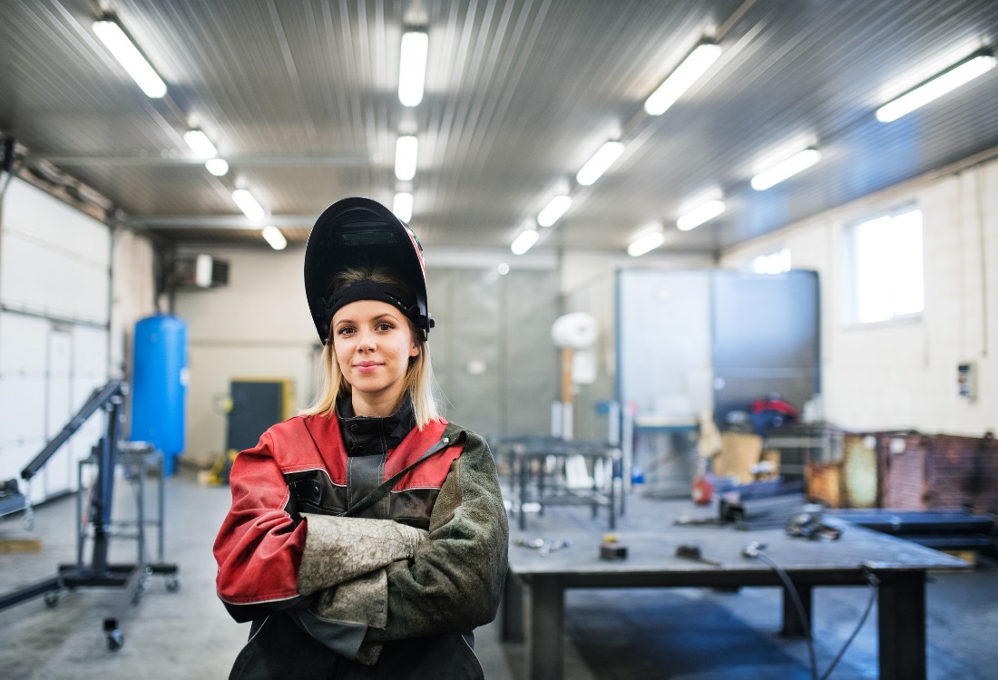 Female metalworker wearing protective welding gear standing confidently in an industrial fabrication workshop. 