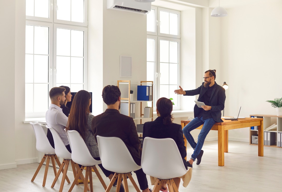 A bearded man with glasses sits on a desk, gesturing to a small group of four people seated in white chairs, appearing to lead a training session that illustrates the importance of recognising and addressing gaps when a management team shows signs it needs training.