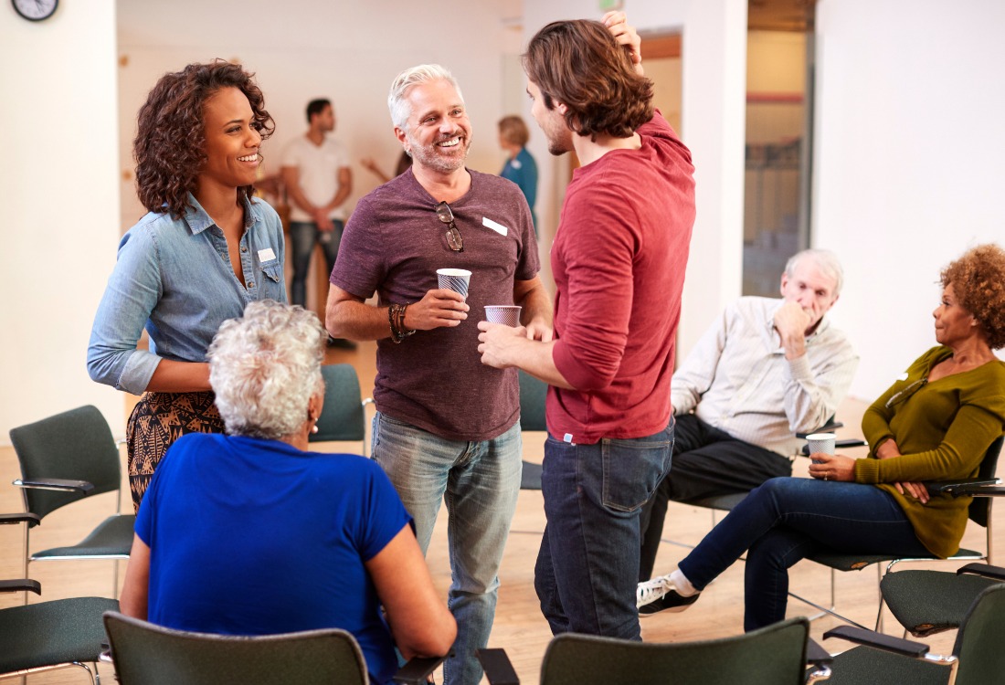 A diverse group of adults gathered in a casual meeting setting, chatting and networking in a circle of chairs, suggesting a community workshop or planning session for local sports or leisure initiatives.