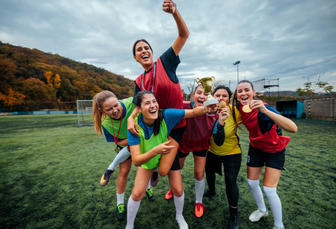 A victorious women's football team celebrates with a trophy and gold medals on the field, embodying the legacy of the Lionesses.