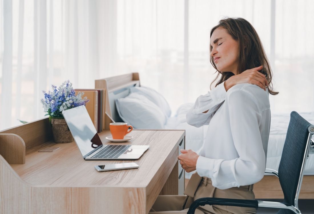 Remote worker sitting at a desk with a laptop, holding her shoulder in discomfort, illustrating the need for ergonomic equipment and proper workstation setup for home-based employees.