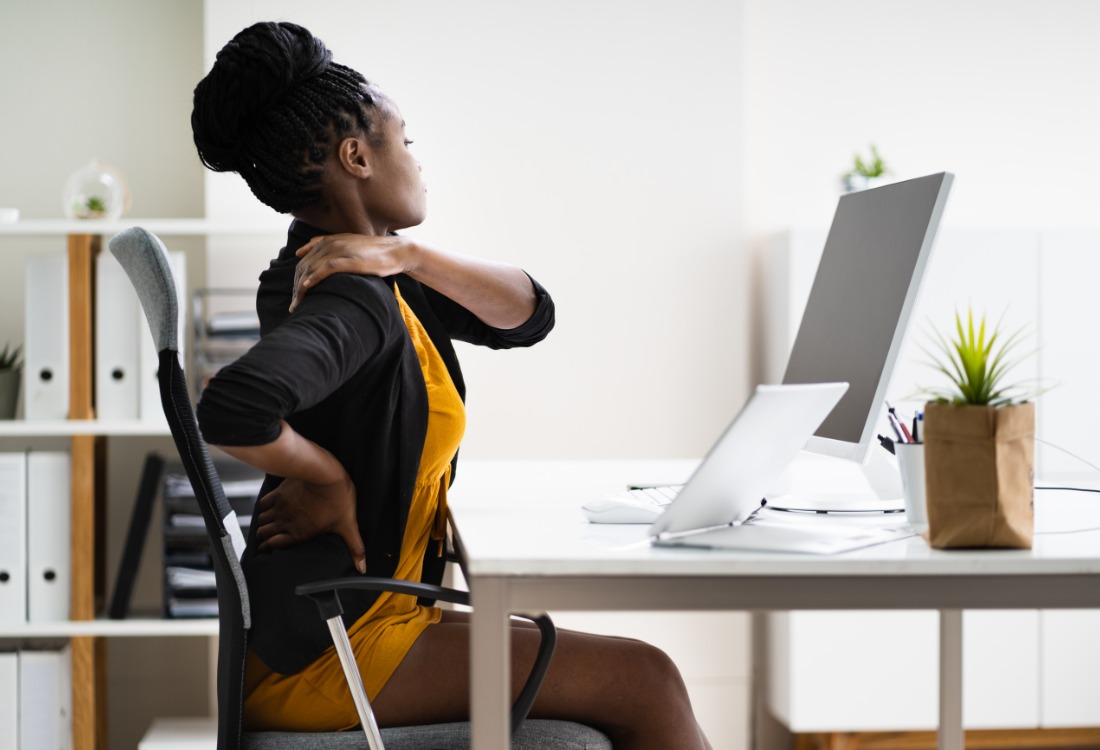 Office worker sitting at a desk holding her back and shoulder in discomfort, illustrating the negative effects of poor workplace ergonomics on employee posture, physical health, and overall wellbeing.