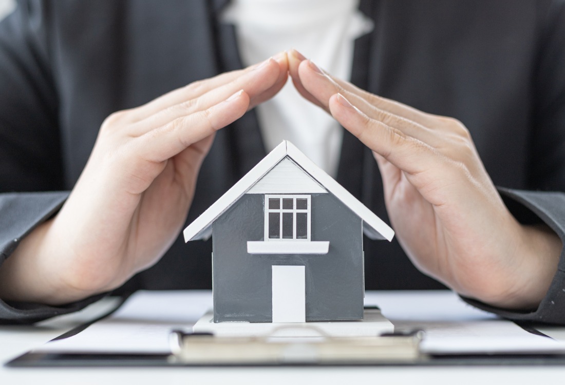 Person shielding a model house with their hands, symbolising insurance for non-standard homes in renovations.