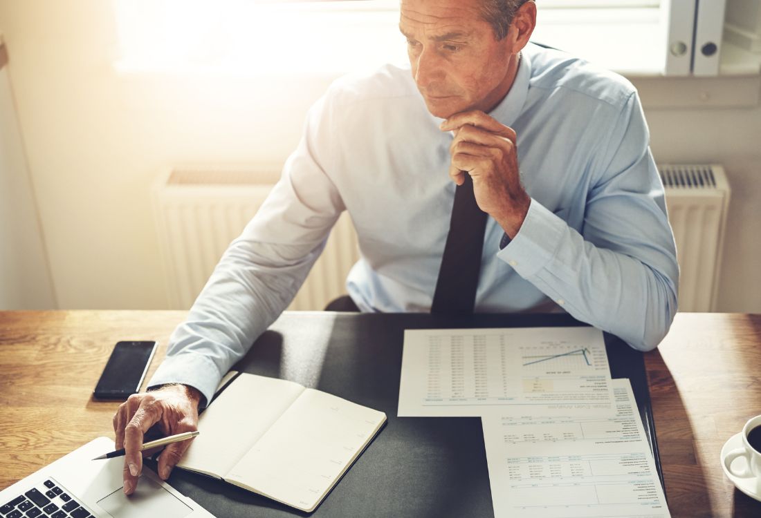 Man working at a desk with spreadsheets, financial reports, and a laptop, reviewing accounting data.