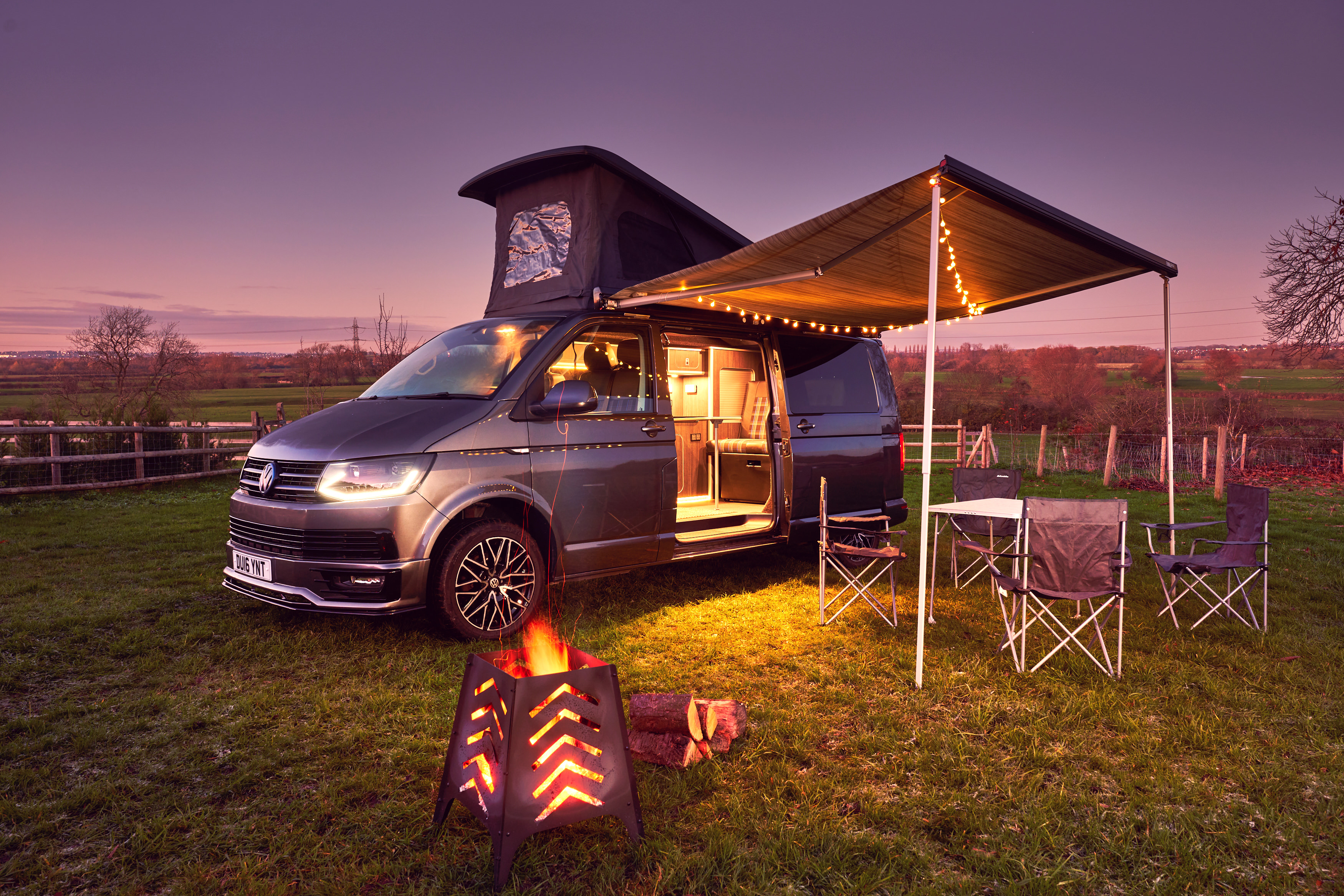 A grey campervan with a pop-up roof and awning is parked in a grassy field at dusk with a fire pit, outdoor furniture, and string lights creating a cosy winter camping scene.