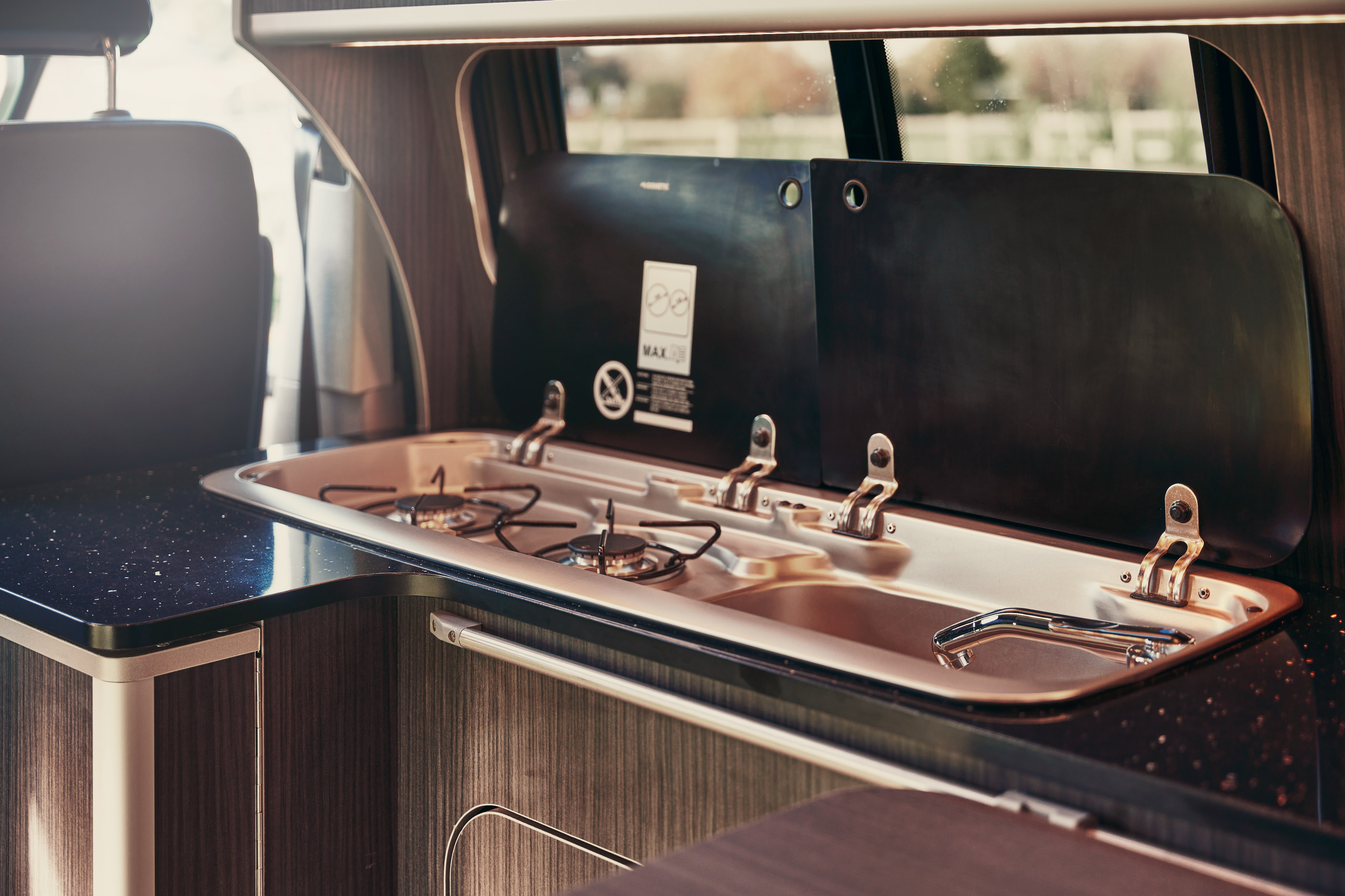 A campervan's compact kitchen with a two-burner gas hob and sink, covered by an open black lid, raising the question of whether its safety checks are similar to those for home gas systems.
