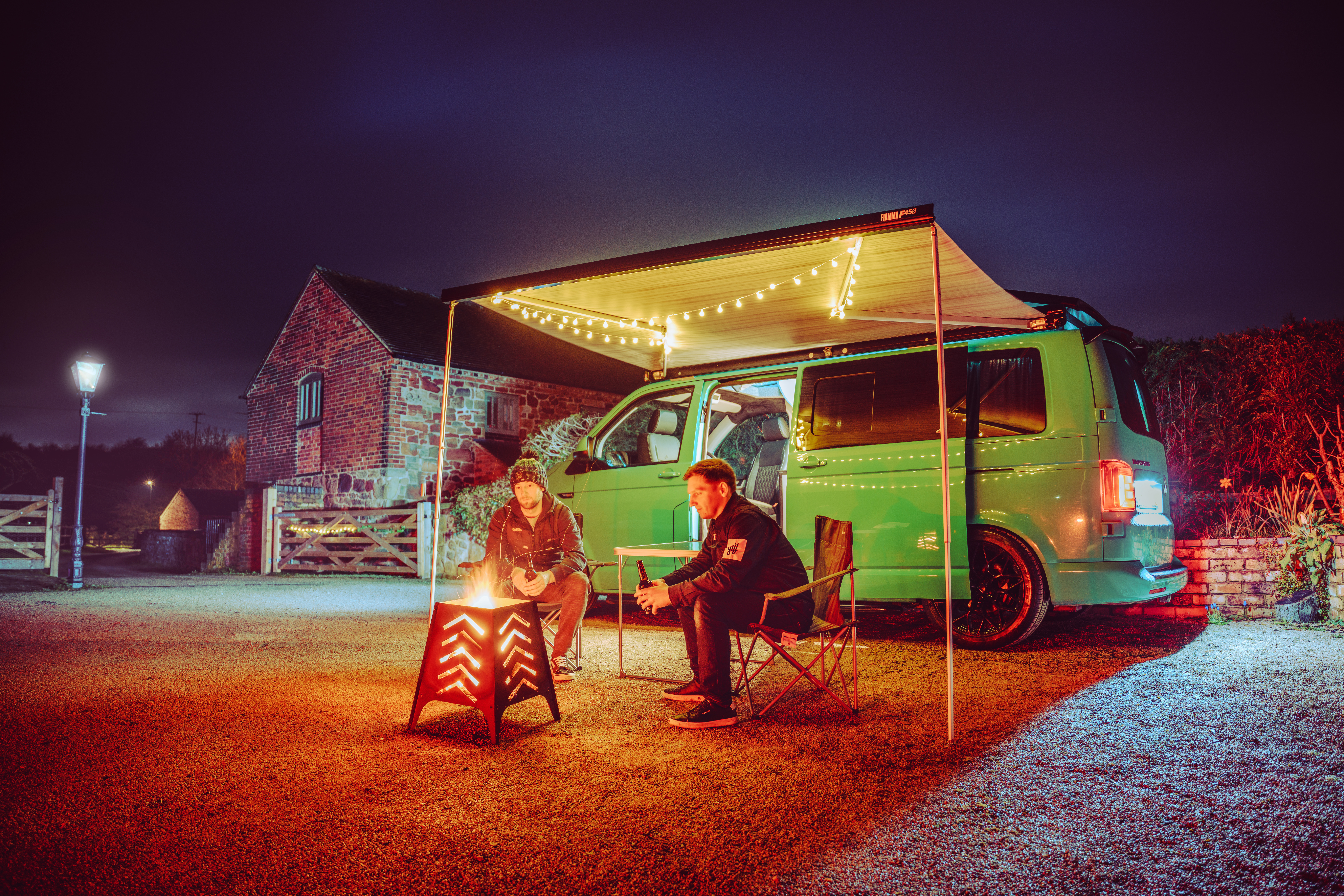 Two men relax by a fire pit under a lit awning attached to a bright green winter campervan at night, with a brick building and a lamppost in the background.